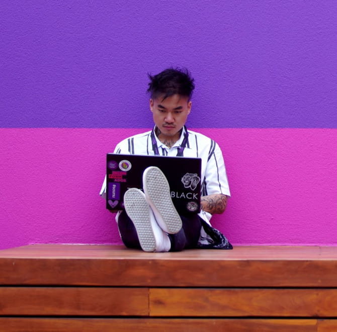 Homem sentado num banco de madeira com suas pernas esticadas e seu notebook no colo. Ele está recostado numa parede rosa e roxa.