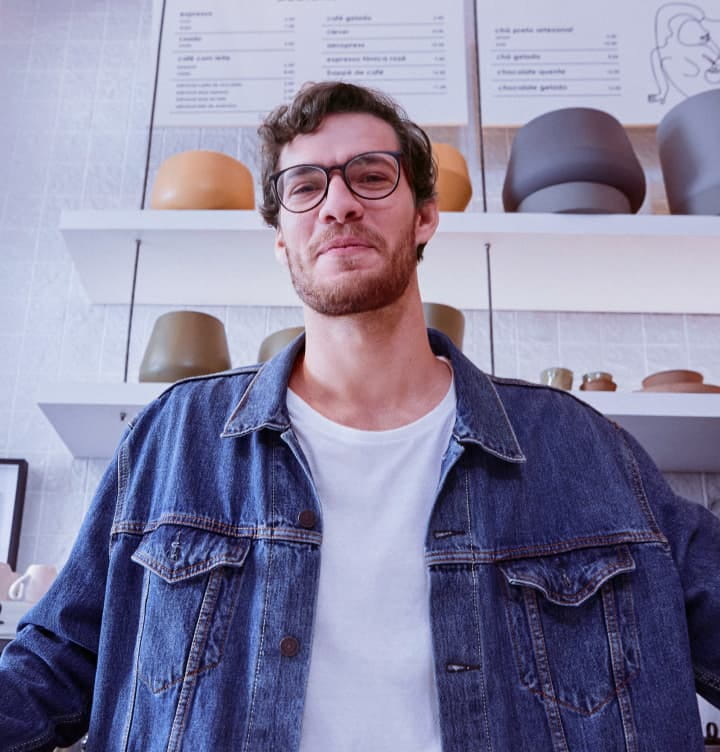 Photography of a man inside a cafe, looking at the camera. He wears glasses, a white T-shirt and a denim jacket. Behind him, two shelves with flowerless vases and an espresso machine. On the left, a window.